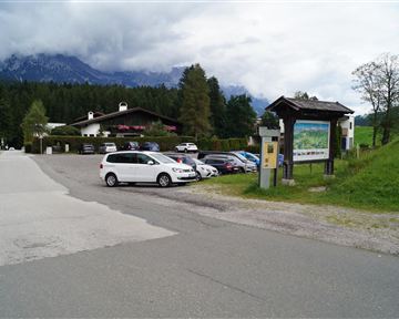 Ein Parkplatz in einer malerischen Umgebung mit Bergen im Hintergrund. Auf der linken Seite ist ein Informationsschild zu sehen.
