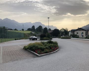Eine ruhige Landschaft mit Bergen im Hintergrund und einer Straße. Zu sehen sind auch einige Blumenbeete und ein parkendes Auto.