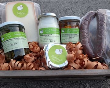 Ein Korb mit verschiedenen Lebensmitteln, darunter Käse, Joghurt und eingelegtes Gemüse. Alles ist hübsch in einem Bett aus Papierstreifen angeordnet.