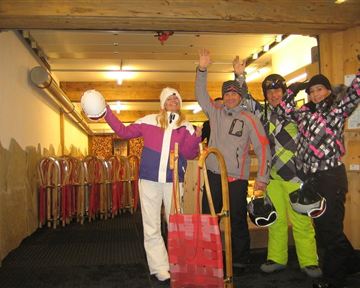 A group of four people in winter clothing stands in a ski room. They are laughing and holding their helmets as well as sleds in their hands.