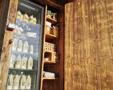 A wooden stand with an open door, offering dairy products and fresh groceries. The refrigerator is visible and displays many bottles in the showcase.