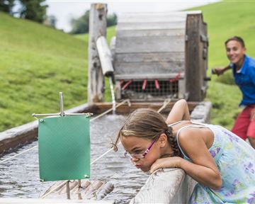 Ein Mädchen beugt sich über eine Wasserrinne und beobachtet das fließende Wasser. Im Hintergrund steht ein Junge und schaut neugierig zu.