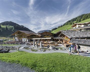 A picturesque alpine inn surrounded by green meadows and gentle hills. Three hikers are walking on a path that leads to the entrance.