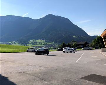 A parking lot with several cars and a view of an impressive mountain landscape. The sky is clear and blue, and the green of the meadows is clearly visible.
