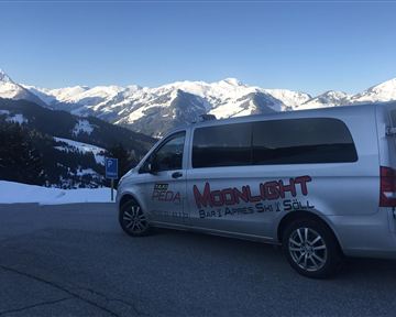 Ein silberner Van steht auf einem Parkplatz mit schneebedeckten Bergen im Hintergrund. Die Umgebung ist winterlich und sonnig.