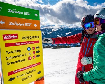 Ein Wintersportler zeigt auf ein Hinweisschild für die SkiWelt Tour in Hopfgarten. Im Hintergrund sind schneebedeckte Berge und ein blauer Himmel zu sehen.