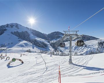 A snowy mountain landscape with a ski lift and bright blue sky. The sun shines brightly over the snow-covered hills.