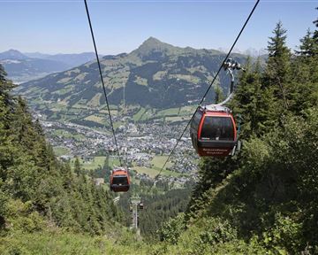 Eine Seilbahn fährt über eine grüne Landschaft mit Blick auf die Berge. Unten erstreckt sich ein malerisches Tal mit kleinen Häusern und viel Natur.