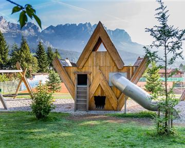 Ein kreativer Spielplatz mit einem hölzernen Spielhaus und einer Rutsche. Im Hintergrund sind grüne Bäume und beeindruckende Berge zu sehen.
