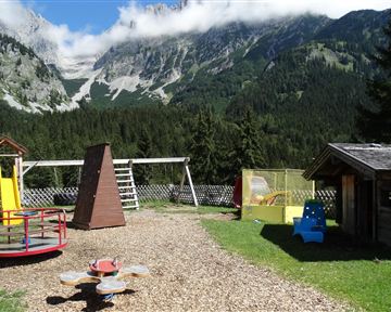 A playground surrounded by mountains and trees. The area features various play equipment and a sunny view.