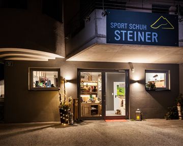 Ein sportlicher Schuhladen mit dem Namen „Sport Schuh Steiner“ ist bei Nacht beleuchtet. Der Eingangsbereich ist einladend, mit dekorativen Elementen an der Fassade.