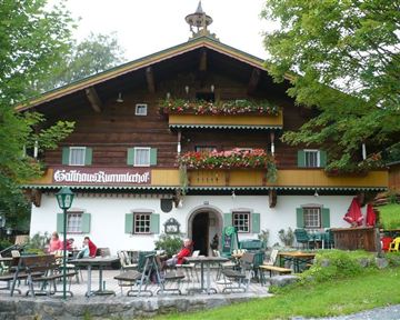 Ein traditionelles Holzhaus mit blühenden Balkonen und einer einladenden Terrasse. Gäste sitzen draußen und genießen die ruhige Umgebung.