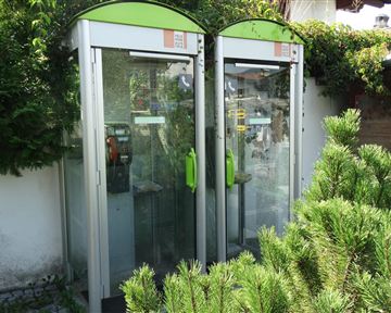Zwei Telefonzellen stehen nebeneinander, umgeben von grünen Pflanzen. Die Telefonzellen haben eine moderne Form und sind teilweise mit Glasfronten ausgestattet.