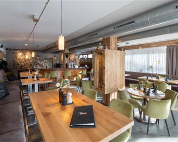 Ein modernes Restaurant mit Holztischen und gemütlichen, grünen Stühlen. Der Raum ist hell und einladend, mit einer Bar im Hintergrund.