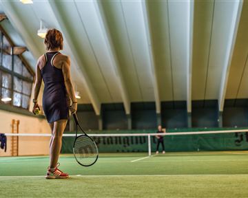 Eine Tennisspielerin steht auf dem Platz und blickt zum Netz. Der Innenbereich ist gut beleuchtet mit einer grünen Spielfläche.