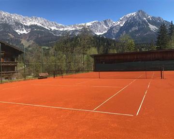 Ein Tennisspielplatz mit rotem Belag, umgeben von majestätischen Bergen und einer klaren blauen Himmel. Die Natur fügt eine malerische Kulisse hinzu.