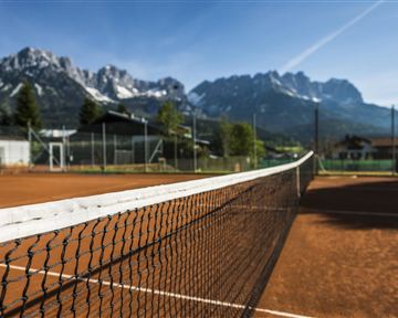 Ein Tennisplatz mit rotem Sand und einem klaren blauen Himmel. Im Hintergrund erheben sich beeindruckende Berge.