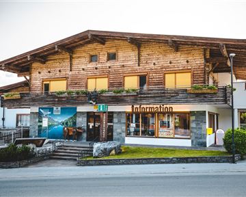 A traditional wooden building with a sign "Information" at the front. It is located on a street and has large windows surrounded by flowers.