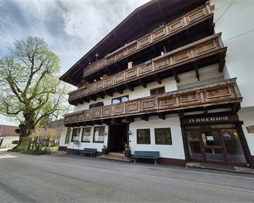 Ein traditionelles alpenländisches Gebäude mit Holzbalustraden und mehreren Etagen. Vor dem Haus befindet sich eine ruhige Straße und ein großer Baum.