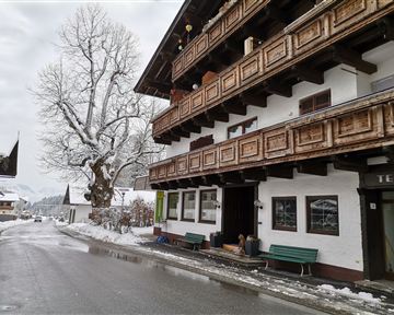 A snowy street with a traditional alpine building. In the background, there is a large tree without leaves.