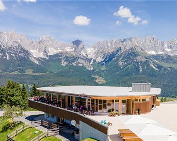 A modern mountain cabin with terrace views of impressive mountains. Clear sky and green forests surround the area.