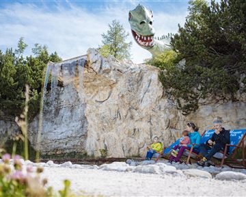 A family sits relaxed on deck chairs in nature. In the background, a huge, fantastical dragon can be seen.