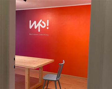 A friendly coworking space with a large table and a chair. The wall is designed in warm colors and features the logo "wp!".