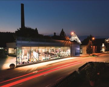 Ein modernes Gebäude mit großen Fenstern, das bei Nacht beleuchtet ist. Im Vordergrund sind Straßenlichter und Pflanzen zu sehen.