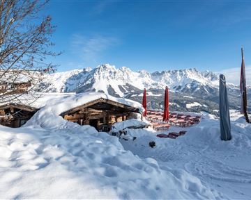 A winter landscape with snowy huts and majestic mountains in the background. The sky is clear and the sun is shining on the snow-covered area.