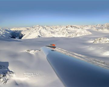 An impressive snow landscape with majestic mountains in the background. In the foreground, a shimmering, reflective wing of an airplane is visible.
