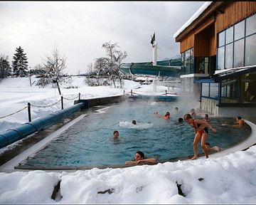 Ein Außenpool in einer winterlichen Umgebung, umgeben von Schnee. Mehrere Personen genießen das warme Wasser und es gibt eine Rutsche neben dem Becken.