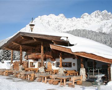 Eine gemütliche Berghütte im Schnee mit Holzterrasse und Tischen. Im Hintergrund sind schneebedeckte Berge zu sehen.
