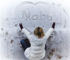 A person is sitting on the snow and writing the name "Noba" in a heart. They are wearing a light gray jacket and red gloves.