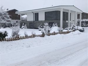 Ein modernes Haus, umgeben von einer weißen Schneedecke. Der Garten ist mit schneebedeckten Pflanzen und einem kleinen Zaun versehen.