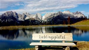 Ein schöner See mit klaren Wasser und Bergen im Hintergrund. Eine Bank mit der Aufschrift "Lieblingsplatzl" lädt zum Entspannen ein.