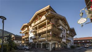 Ein modernes Gebäude im alpinen Stil mit Balkonen und Blumen. Es liegt in einer ruhigen Straße mit bergen im Hintergrund.