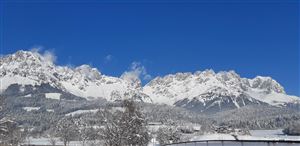 An impressive mountain landscape with snow-covered peaks under a clear blue sky. In the foreground, snow-covered trees can be seen.