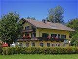 A yellow house with a balcony, adorned with flowers. It stands surrounded by trees and a well-maintained garden.