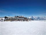 A mountainous winter landscape with plenty of snow and a clear blue sky. In the foreground stands a ski lodge, surrounded by skiers.