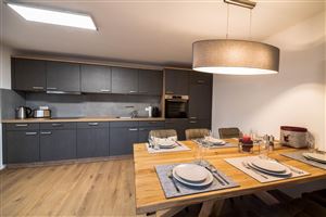 Eine moderne Küche mit dunklen Schränken und einem großen Esstisch. Der Tisch ist festlich gedeckt und eine helle Deckenlampe sorgt für gutes Licht.