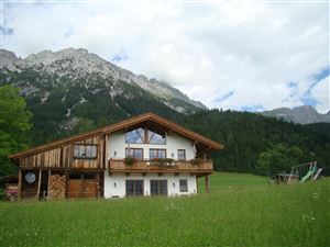 Ein schönes, traditionelles chaletartiges Haus in den Alpen. Im Hintergrund sind majestätische Berge und ein grünes Feld zu sehen.