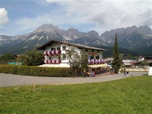 A picturesque house with blooming plants and a garden. In the background, majestic mountains rise beneath a partly cloudy sky.