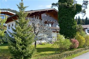 Ein schönes Holzhaus umgeben von Bäumen und Blumen. Der Himmel ist blau und die Landschaft wirkt einladend und grün.