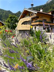Ein schöner Bauernhof mit dem Namen „Hof Alberts“ ist von bunten Blumen und Lavendel umgeben. Im Hintergrund sind Berge und ein blaues Himmel sichtbar.