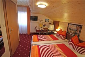 Ein gemütliches Schlafzimmer mit zwei Betten und bunten Kissen. Es gibt einen kleinen Tisch, einen Fernseher und viel Tageslicht durch das Fenster.