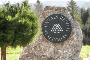 A rock with a sign displaying "Mountain Resort Seiwald." In the background, green trees and a picturesque mountain landscape can be seen.