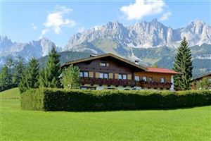 Ein traditionelles Haus umgeben von einer grünen Wiese und Bäumen. Im Hintergrund erheben sich majestätische Berge unter einem blauen Himmel.