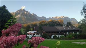 Eine malerische Berglandschaft mit hohen Gipfeln im Hintergrund. Im Vordergrund blühen pinke Blumen vor einem gemütlichen Gebäude.
