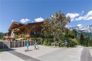 Ein schönes Holzhaus umgeben von Bäumen und Bergen. Kinder fahren mit ihren Rollern auf einem ruhigen Weg.
