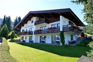Ein charmantes Haus mit Balkonen, umgeben von einer grünen Wiese und hohen Bäumen. Die Architektur ist typisch für alpine Regionen.